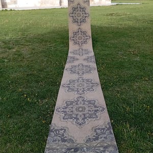 Può includere: Un tappeto passatoia lungo e stretto con uno sfondo beige chiaro e un motivo ripetuto di medaglioni floreali grigio scuro. Il tappeto presenta un bordo decorativo all'estremità con intricati motivi geometrici e botanici.