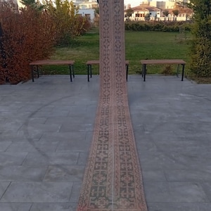 May include: A long, rectangular rug with a detailed geometric pattern in shades of brown, green, and red. The rug is displayed outdoors, showcasing its length and intricate design. The rug's edges have a fringed border.