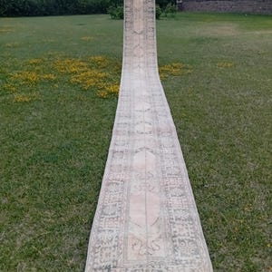 May include: An exceptionally long, narrow runner rug featuring a faded, muted color palette of beige, cream, and soft grey. The rug displays a repeating geometric and floral pattern throughout its length, framed by a detailed, intricate border.