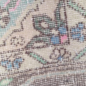 May include: A woven textile rug featuring a detailed geometric and floral pattern in shades of beige, dark brown, sage green, light blue, and small accents of pink. The design consists of repeating motifs with dark brown outlines on a beige background.