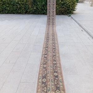 May include: A long, narrow runner rug with a detailed pattern. The rug features a central design with repeating geometric shapes in shades of brown, beige, and blue. The rug is displayed outdoors, showcasing its length and intricate design.