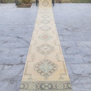 May include: A long, rectangular beige runner rug with an intricate geometric pattern in shades of green, brown, and red. The rug is placed on a grey stone surface, showcasing its length and design.