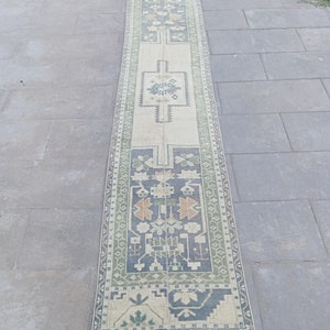 Op de afbeelding: Een lang, smal tapijt met een crèmekleurige achtergrond en een geometrisch patroon in tinten groen, blauw en oranje. Het tapijt heeft aan beide uiteinden franjes.