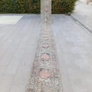 May include: A long, narrow, vintage runner rug with a faded, distressed appearance. The rug features a repeating pattern of pink and beige medallions on a muted green and brown background. The rug is displayed outdoors.