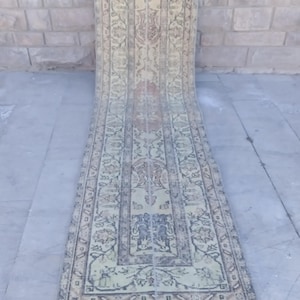 Pode incluir: Um tapete corredor antigo com uma aparência desbotada e desgastada. O tapete apresenta um design central com padrões florais e geométricos em tons de bege, verde e vermelho suave. Está estendido sobre uma superfície de concreto.