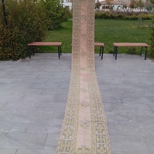 May include: A long, rectangular rug in faded pink and beige. It has an intricate geometric pattern with floral accents. The rug is displayed outdoors, standing upright against a backdrop of greenery and benches. The rug's length is visible.