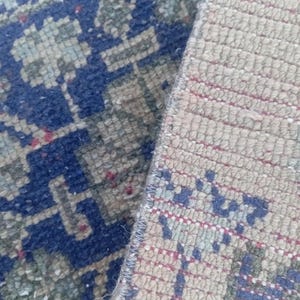 May include: A woven rug featuring a deep blue section with light green and red floral motifs alongside a beige section with similar patterns. The rug displays a dense, knotted pile texture with visible edges and a distinct color contrast between the two areas.