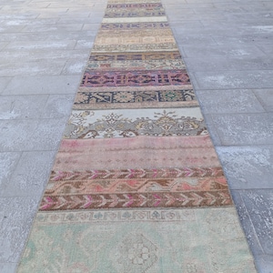 Puede incluir: Una alfombra larga y estrecha hecha de varias tiras de tela de colores. La alfombra tiene un diseño de patchwork con diferentes patrones y colores, incluyendo rosa, marrón, beige y verde. La alfombra está colocada sobre un suelo de baldosas grises.