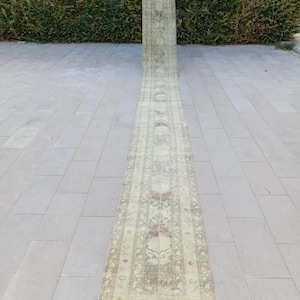 Peut inclure: Un long tapis rectangulaire vintage à l'aspect délavé. Le tapis présente un motif floral central dans des tons beige, marron et vert sur un fond clair. Le tapis est enroulé à une extrémité et déroulé sur une surface en pierre.