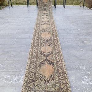 May include: A long, rectangular runner rug with an intricate pattern in brown, beige, and olive green. The rug has a central design with geometric shapes and floral motifs. The rug is displayed outdoors, likely on a patio or walkway.