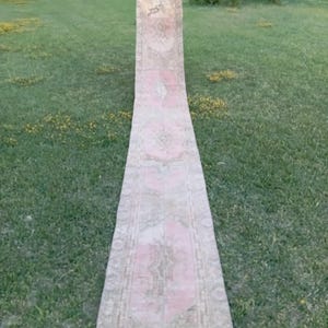 Könnte beinhalten: Ein langer, schmaler Läuferteppich mit einer verblassten, gedämpften Farbpalette aus zartem Rosa, Beige und Creme. Der Teppich zeigt ein sich wiederholendes geometrisches und florales Medaillonmuster über die gesamte Länge mit einer Bordüre.