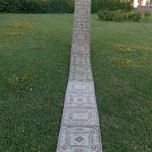 May include: A long, narrow runner rug featuring a repeating geometric pattern in shades of beige, grey, and muted brown. The rug displays a series of interconnected octagonal motifs with intricate linear details and a fringed edge at the base.