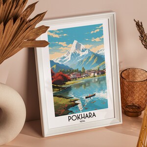 May include: A white framed print of a vintage travel poster for Pokhara, Nepal. The poster features a colourful illustration of a mountain range, a lake, and a small village. The text "POKHARA NEPAL" is printed at the bottom of the poster.