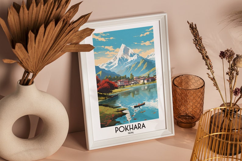 May include: A white framed print of a vintage travel poster for Pokhara, Nepal. The poster features a colourful illustration of a mountain range, a lake, and a small village. The text "POKHARA NEPAL" is printed at the bottom of the poster.