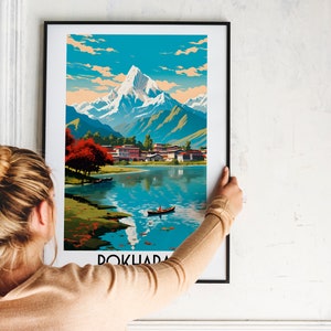 May include: A framed poster featuring a vintage travel illustration of a mountain range, a lake, and a small village. The poster is titled "Pokhara" and is in a black frame.