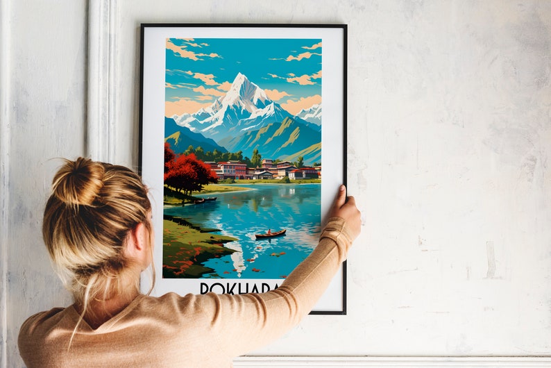 May include: A framed poster featuring a vintage travel illustration of a mountain range, a lake, and a small village. The poster is titled "Pokhara" and is in a black frame.