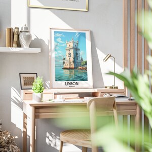 Op de afbeelding: Een ingelijste poster met een vintage reisillustratie van de Toren van Bel&eacute;m in Lissabon, Portugal. De poster hangt aan een witte muur boven een houten bureau met een stoel en een lamp.