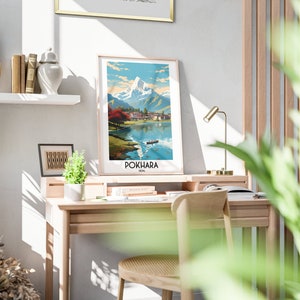 May include: A framed print of a mountain landscape with a lake in the foreground. The print is titled "POKHARA, NEPAL". The print is hanging on a white wall above a wooden desk with a chair and a lamp.