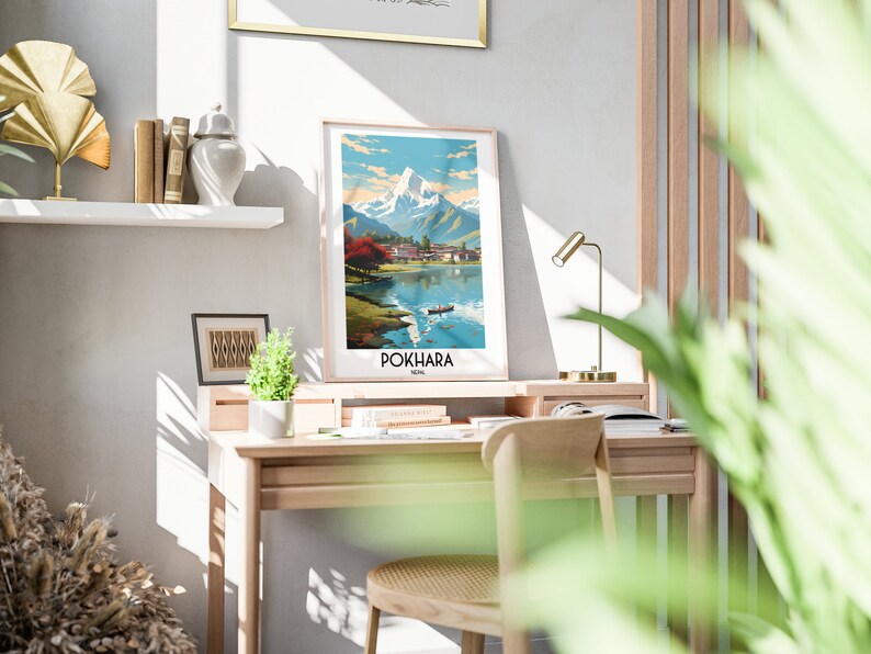 May include: A framed print of a mountain landscape with a lake in the foreground. The print is titled "POKHARA, NEPAL". The print is hanging on a white wall above a wooden desk with a chair and a lamp.