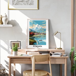 May include: A vintage travel poster featuring a colourful illustration of a mountain range and a lake with a boat in the foreground. The poster is framed and hanging on a wall above a wooden desk. The text on the poster reads "POKHARA NEPAL".