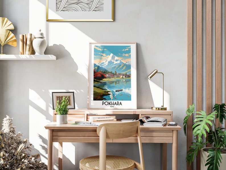 May include: A vintage travel poster featuring a colourful illustration of a mountain range and a lake with a boat in the foreground. The poster is framed and hanging on a wall above a wooden desk. The text on the poster reads "POKHARA NEPAL".