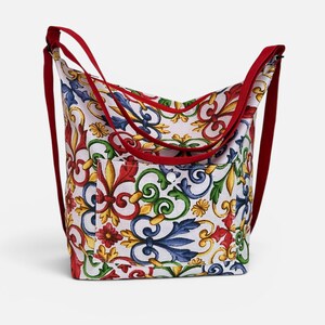 May include: A hobo-style shoulder bag featuring a white base with a vibrant, multi-colored baroque-inspired floral and scroll pattern in red, blue, green, and yellow. The bag includes a front pocket with a snap closure and solid red straps.