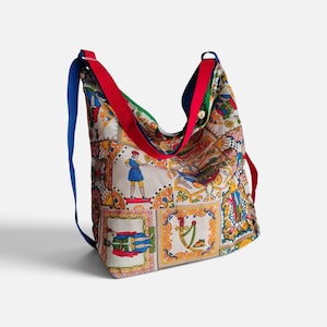 May include: A hobo bag featuring a multi-colored patchwork print of historical figures and ornate decorative borders. The bag has a wide red shoulder strap and a thin blue strap, with a light-colored fabric base and various colorful illustrations.