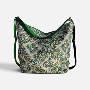 May include: A hobo-style shoulder bag featuring a white base with a repeating green and yellow ornate tile-inspired pattern. The bag includes a solid green interior lining and a wide black shoulder strap attached to the sides of the bag.