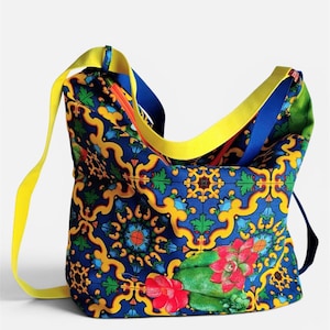 May include: A hobo bag featuring a vibrant blue background with a repeating yellow and orange scrollwork pattern, accented by green foliage and red flowers. The bag includes a bright yellow shoulder strap and a contrasting blue interior lining.