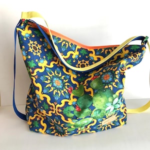 Puede incluir: Bolso de hombro azul con un vibrante estampado de diseños florales amarillos y verdes y una ilustración de cactus. El bolso tiene una correa amarilla y una correa azul, con una cremallera naranja.