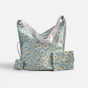 May include: A light blue hobo bag featuring a white daisy pattern with yellow centers. The bag includes a wide white shoulder strap, a small attached pouch on a cord, and a separate rectangular pocket with a white snap button closure on the front.