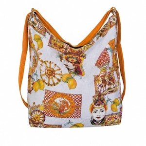 May include: A hobo-style shoulder bag featuring a white base with a vibrant, multi-colored print of lemons, ornate wheels, and stylized portraits of people wearing headpieces. The bag includes two solid orange straps attached to the top corners.