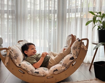 May include: A child sits on a wooden rocking chair with a padded cushion featuring a white background with black and gray animal illustrations. The rocking chair is made of natural wood and has a curved shape. The child is wearing a green shirt and beige pants.