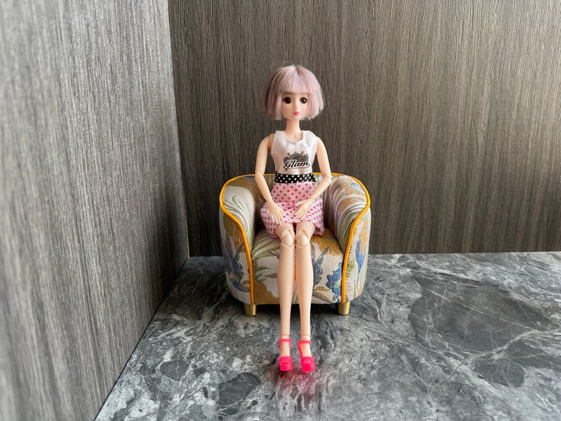 May include: A fashion doll with short, light purple hair sits in a miniature armchair. The doll wears a white top with the word "Glam", a pink skirt with white polka dots, and bright pink heels. The armchair has a floral pattern.