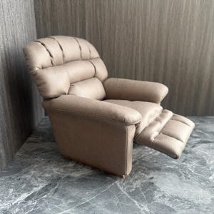 Könnte beinhalten: Ein Miniatur-Liegesessel in Beige mit gepolsterter Kopfstütze, Armlehnen und Fußstütze. Der Sessel ist aus einem weichen Stoff gefertigt und hat eine Holzbasis. Die Fußstütze ist ausgezogen, und der Stuhl steht vor einem grauen Hintergrund.