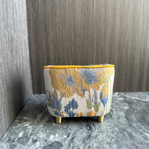 May include: A small, upholstered ottoman with a floral pattern in yellow, blue, and green on a cream background. The top edge is trimmed with yellow piping, and it has short, gold-colored legs. The ottoman is a decorative furniture piece.