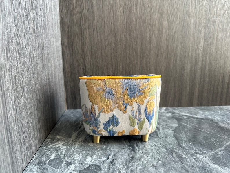 May include: A small, upholstered ottoman with a floral pattern in yellow, blue, and green on a cream background. The top edge is trimmed with yellow piping, and it has short, gold-colored legs. The ottoman is a decorative furniture piece.