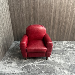 May include: A small, red leather armchair with a tufted back and rolled arms. The chair is sitting on a gray marble floor.