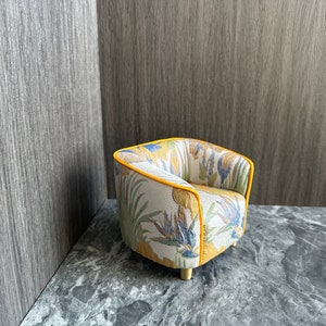 May include: A small, decorative armchair with a botanical print featuring yellow, blue, and green flowers and leaves on a cream background. The chair has a yellow trim and gold-colored legs. The chair is set against a gray background.