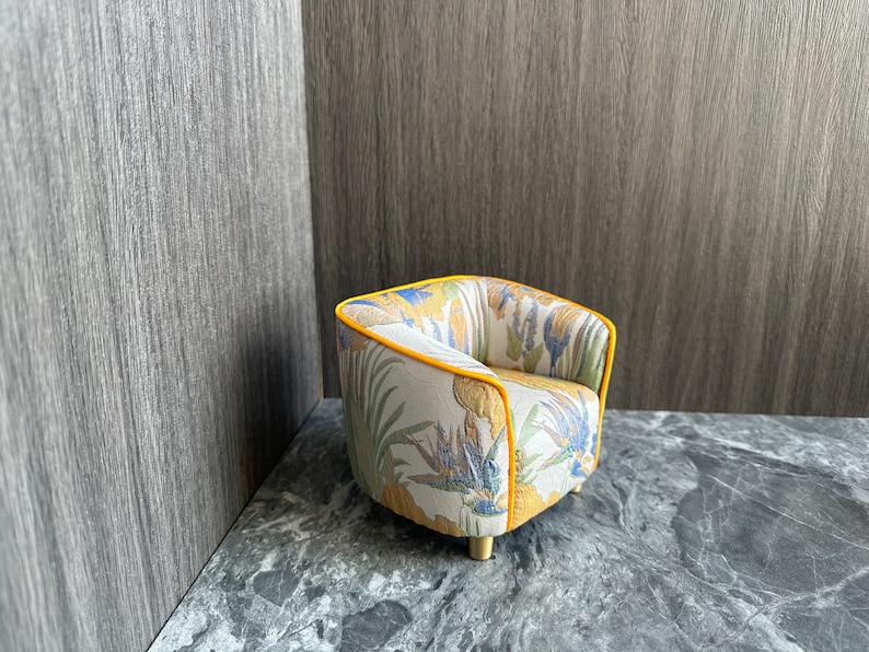 May include: A small, decorative armchair with a botanical print featuring yellow, blue, and green flowers and leaves on a cream background. The chair has a yellow trim and gold-colored legs. The chair is set against a gray background.