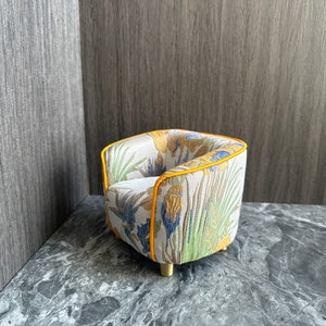 May include: A miniature armchair with a floral pattern in yellow, blue, and green on a cream background. The chair has a yellow trim and gold-colored legs. The overall design is a small-scale furniture piece.