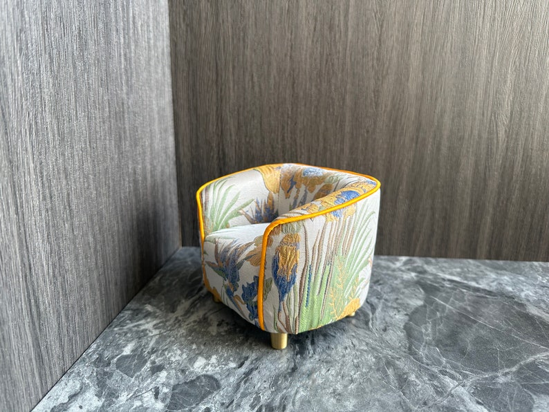 May include: A miniature armchair with a floral pattern in yellow, blue, and green on a cream background. The chair has a yellow trim and gold-colored legs. The overall design is a small-scale furniture piece.
