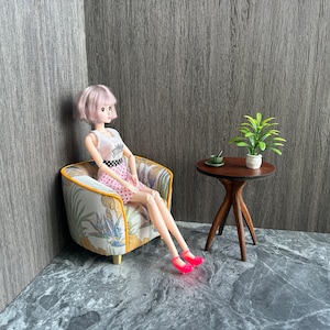 May include: A doll in a pink polka dot skirt and white top sits in a miniature armchair with a floral pattern. A small wooden table holds a plant and a cup. The doll wears pink shoes.
