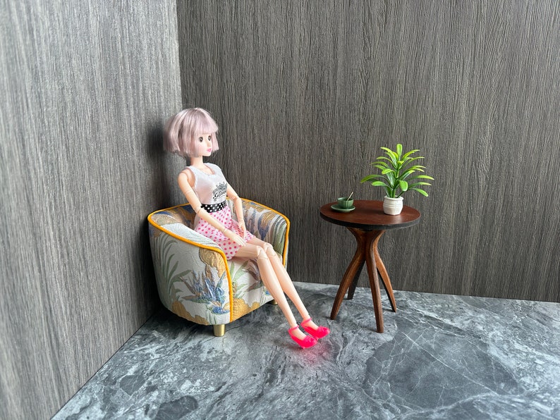 May include: A doll in a pink polka dot skirt and white top sits in a miniature armchair with a floral pattern. A small wooden table holds a plant and a cup. The doll wears pink shoes.