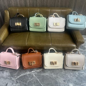 May include: A collection of miniature handbags in various colors, including black, green, white, and pink. Each bag features a top handle, a gold-colored clasp, and a structured design. The bags are arranged on a green leather couch and a marble surface.
