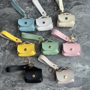 May include: A collection of small, colorful leather pouches with attached wrist straps and gold-tone hardware. The pouches are in various pastel colors, including blue, white, yellow, green, pink, black, and beige. Each pouch has a small, round button closure.