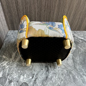 May include: Upholstered ottoman with a floral pattern in blue, gold, and white. The edges are trimmed with yellow piping. It has four brass-colored legs and a black bottom. A decorative furniture piece.