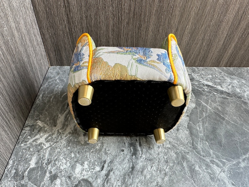 May include: Upholstered ottoman with a floral pattern in blue, gold, and white. The edges are trimmed with yellow piping. It has four brass-colored legs and a black bottom. A decorative furniture piece.