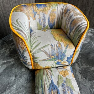 May include: A small, upholstered armchair and footstool set. The chair and footstool feature a floral pattern in shades of blue, green, and gold on a cream background. The edges of the chair are trimmed with a bright yellow piping.