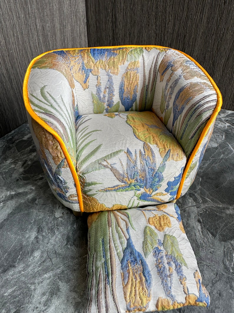 May include: A small, upholstered armchair and footstool set. The chair and footstool feature a floral pattern in shades of blue, green, and gold on a cream background. The edges of the chair are trimmed with a bright yellow piping.